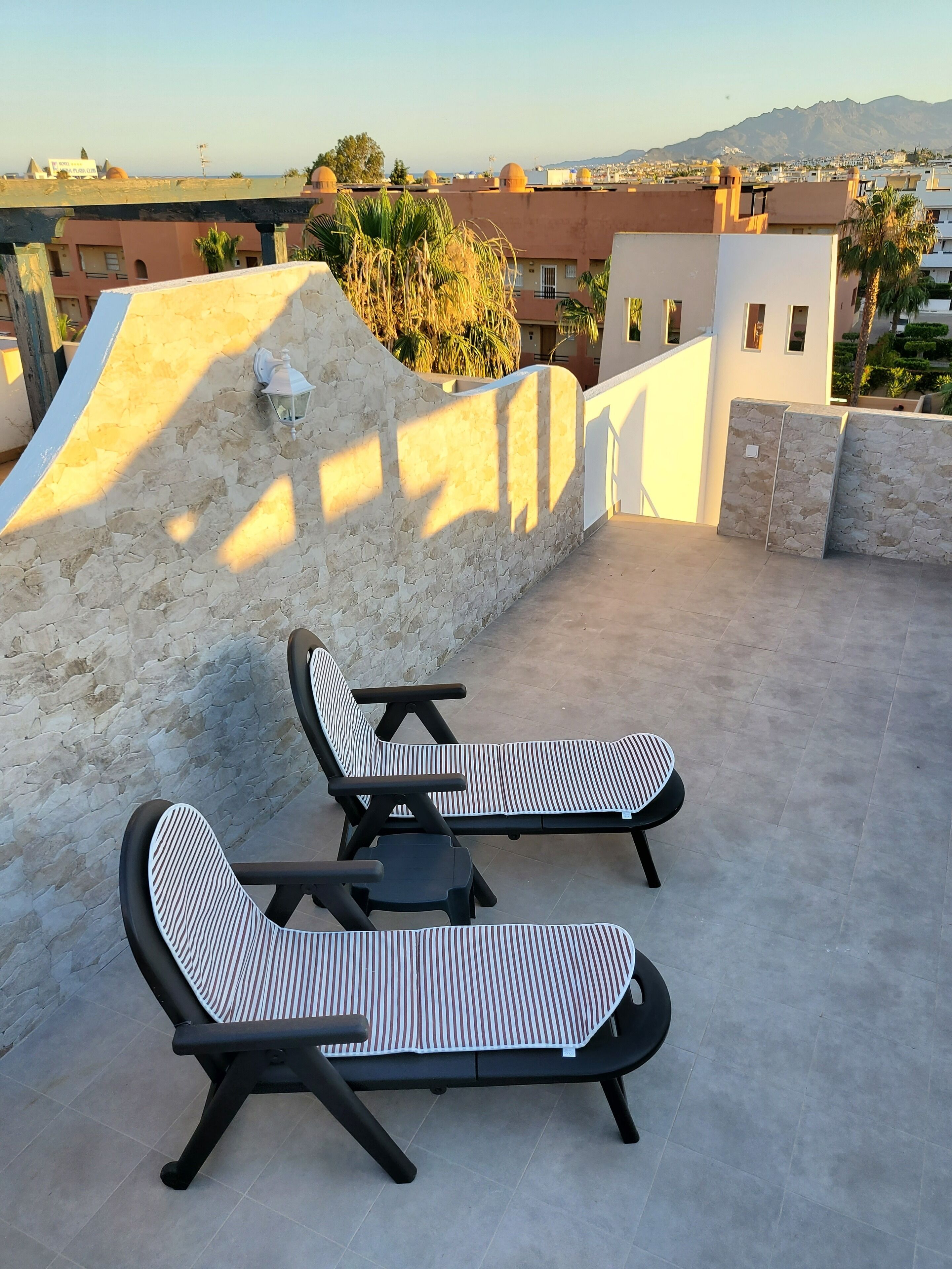 Terraza en la azotea con dos sillones y una mesa, con vistas a un paisaje urbano y montañas.