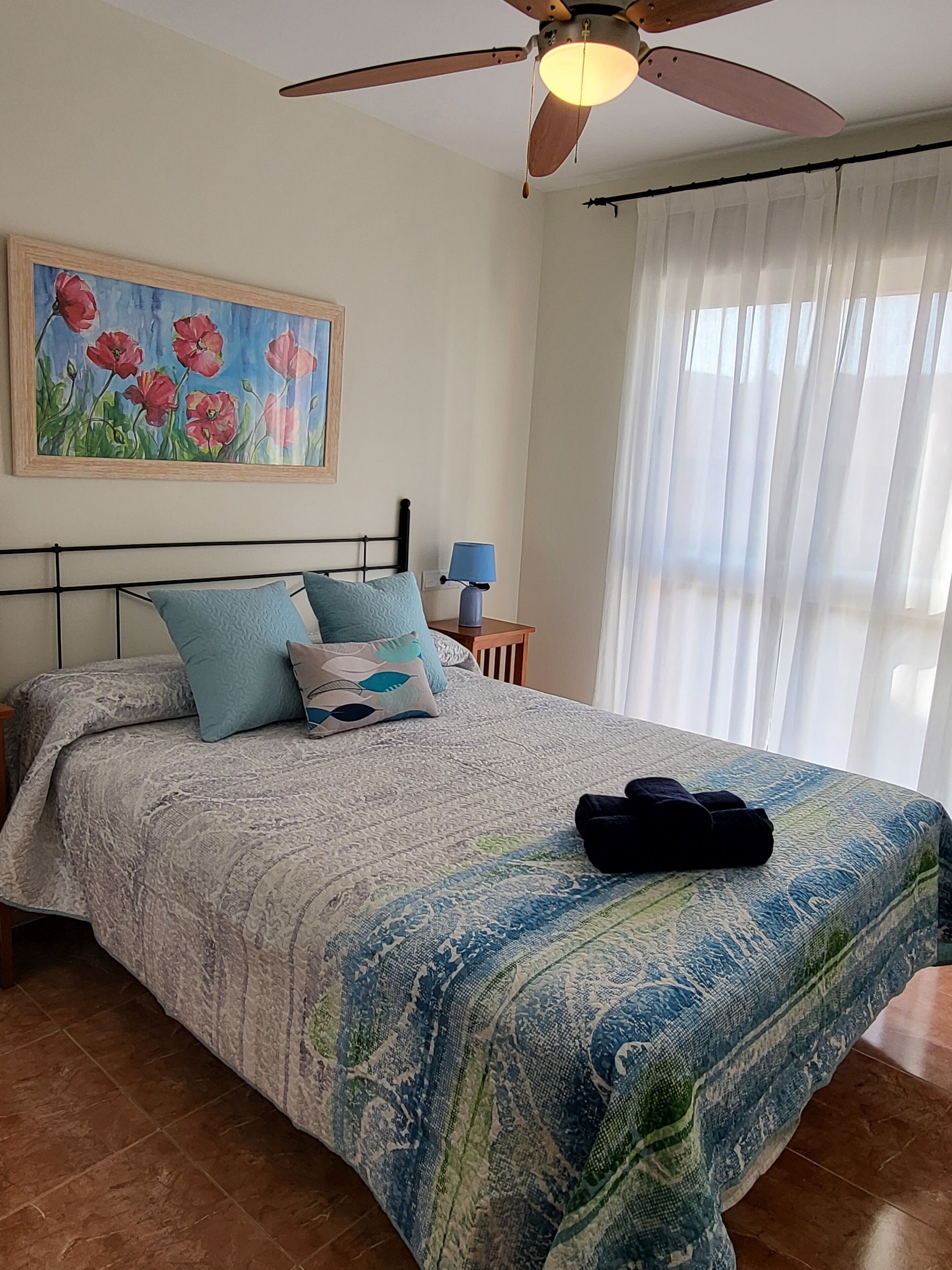Un dormitorio con cama, ventilador de techo, un cuadro, una mesita de noche y una ventana con cortinas.