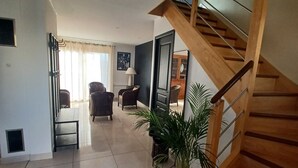 Una sala de estar moderna con una escalera de madera, una zona de comedor con sillas y una planta grande en maceta.