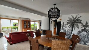 Un área de comedor con una mesa redonda y sillas de mimbre, un sofá rojo y un ventanal con vistas a la vegetación.