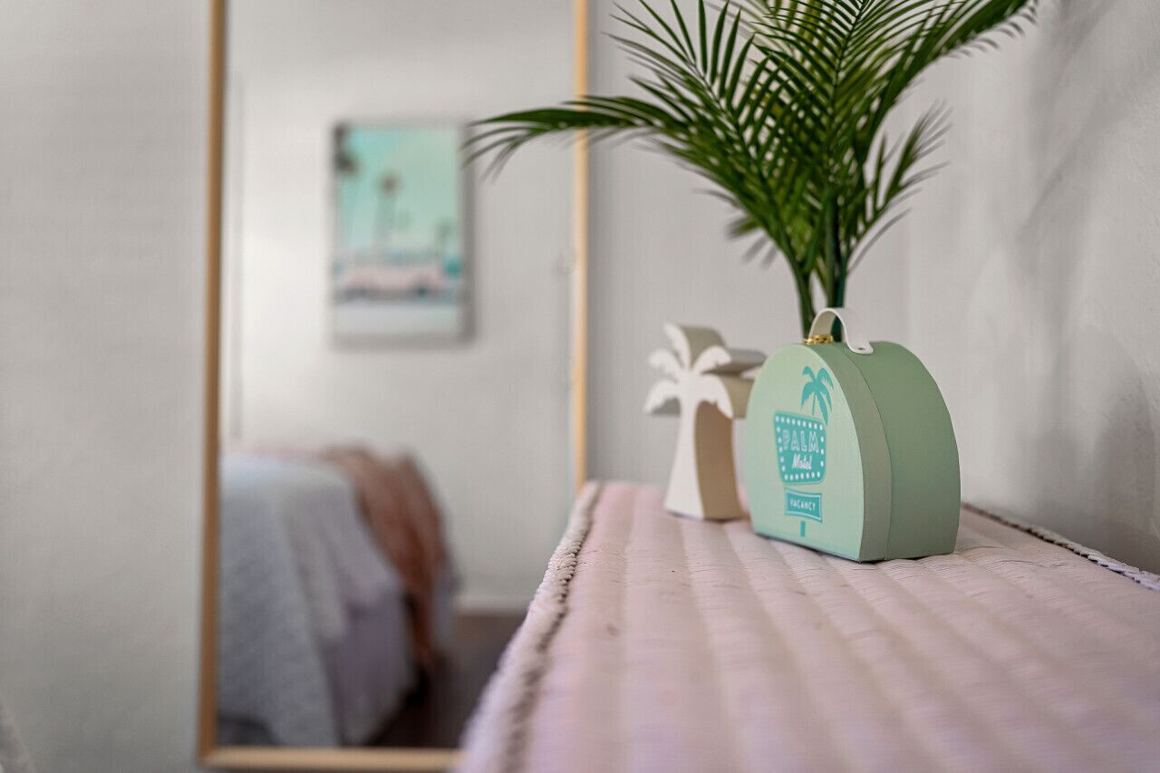 Een kamer met een bed, een spiegel, een kleine plant in een decoratieve pot en een ingelijst schilderij aan de muur.
