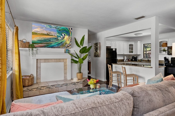 A modern living room with a sofa, a coffee table, and a fireplace. There is a kitchen area with white cabinets and a refrigerator.