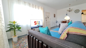 Una sala de estar con un sofá, un televisor mostrando 'Super Simple Songs', una planta en maceta y un tapete estampado.