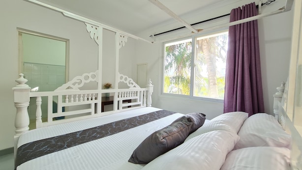 Un dormitorio con cama blanca, ventana con vista a la vegetación y cortina morada.