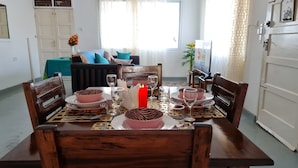 Un comedor con una mesa de madera dispuesta para cuatro personas, rodeada de sillas. Se observa un sofá y un televisor al fondo.
