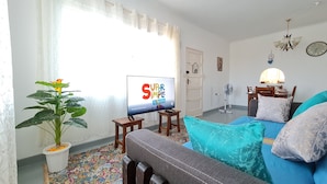 Un salón con una televisión mostrando 'Super Simple Songs', una planta en maceta, un sofá con cojines, una mesa de comedor con sillas y un ventilador.