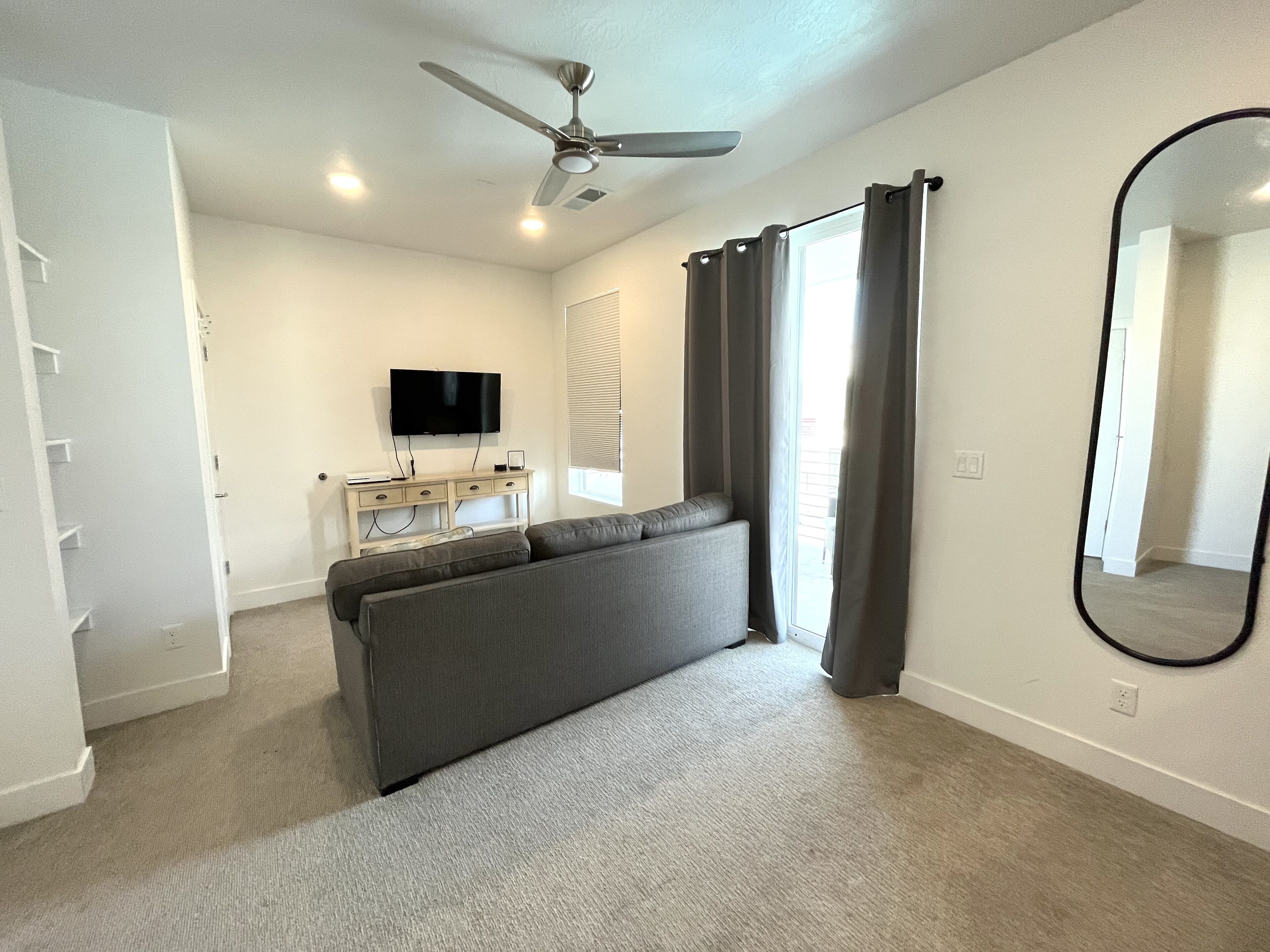 A modern, well-lit room with a ceiling fan, a flat-screen TV, a grey sofa, a small white console table, and a large mirror.