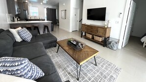 Un salon moderne avec un canapé d’angle gris, une table basse en bois et un téléviseur posé sur un meuble en bois. On y trouve également un coin cuisine avec un bar et des tabourets, ainsi qu’un escalier menant à un autre niveau.
