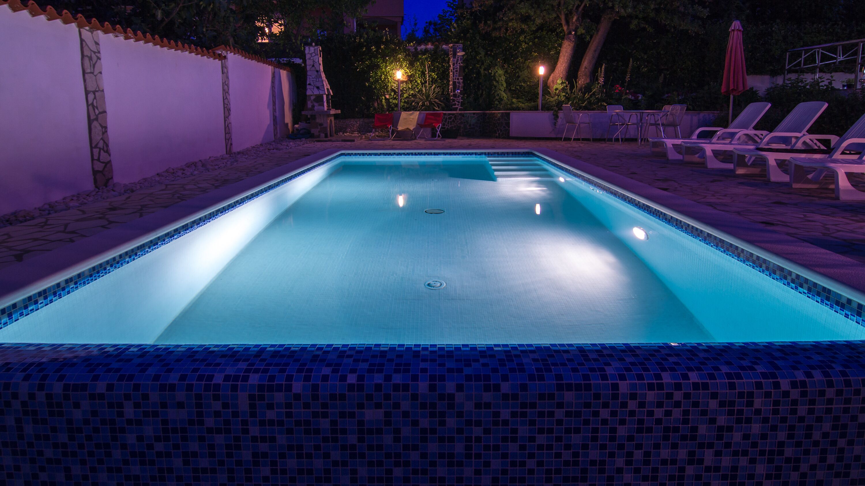 A rectangular swimming pool with a tiled edge, surrounded by lounge chairs and a stone wall.