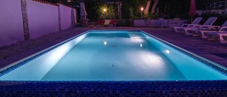 A rectangular swimming pool with a tiled edge, surrounded by lounge chairs and a stone wall.
