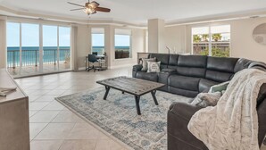 A spacious living room with a large window overlooking the ocean, a ceiling fan, and a black leather sofa.