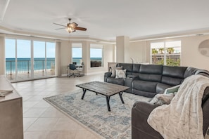 A spacious living room with a large window overlooking the ocean, a ceiling fan, and a black leather sofa.