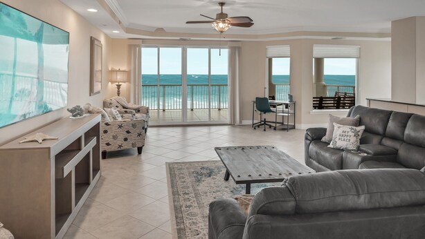 A spacious living room with a large TV, comfortable seating, a ceiling fan, and a view of the ocean through large windows.