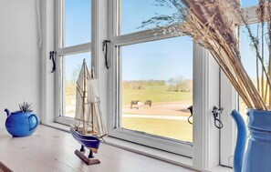 Ein Fenster mit Blick auf ein grasbewachsenes Feld und Pferde, ein kleines Segelbootmodell und zwei Vasen auf einer Holzablage.