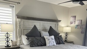 Un dormitorio con una cama grande, dos mesitas de noche, un ventilador de techo y una ventana con persianas.