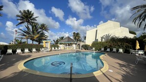 Piscina con sillas de descanso y sombrillas, rodeada de palmeras y un edificio con fachada blanca.