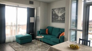 A modern living room with a teal sofa, a small round table, and a dining table with fruit.