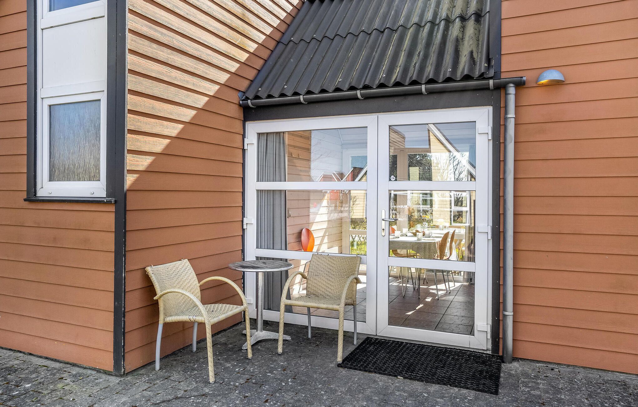 Un coin d’un bâtiment avec une porte-fenêtre coulissante donnant sur un espace extérieur aménagé avec deux fauteuils en osier et une petite table.