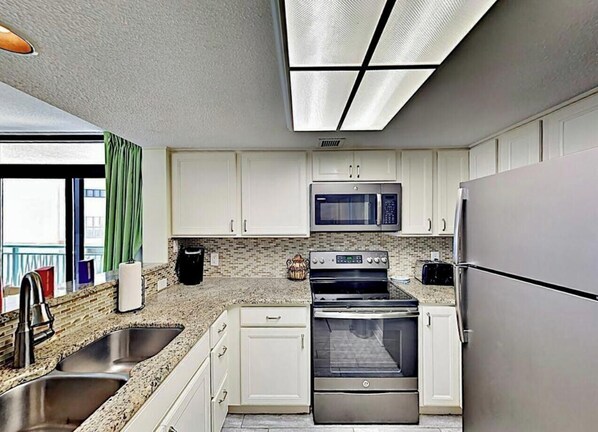 A modern kitchen with stainless steel appliances, a double sink, and a view of the balcony. - Endless Ocean Abode - Direct Ocean views (Myrtle Beach)
