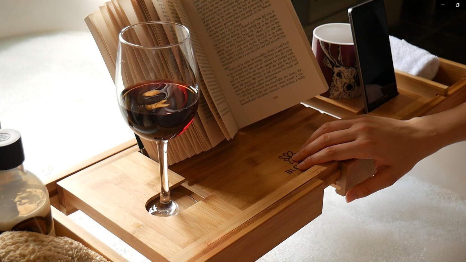 Ein Holztablett mit einem Weinglas, einem offenen Buch, einem Smartphone und einer Tasse auf einem Tisch.