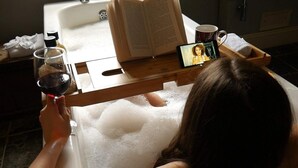 Une personne se détend dans un bain, un livre à la main, avec un téléphone intelligent et un verre de vin à portée de main.