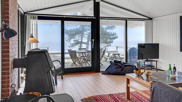 Ein modernes Wohnzimmer mit einer großen Glasschiebetür, die auf eine Terrasse im Freien führt. Im Raum befindet sich ein schwarzer Sessel, ein kleiner Tisch mit einer Lampe und ein Fernseher auf einem Schrank.