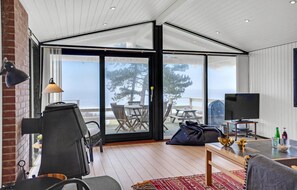 Een moderne woonkamer met een grote schuifpui die uitkomt op een terras. Binnen is een zwarte fauteuil, een kleine tafel met een lamp en een televisie op een dressoir.