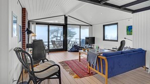 Ein großzügiger Wohnbereich mit blauem Sofa, Esstisch und Fernseher. Ein großes Fenster bietet Blick ins Freie.