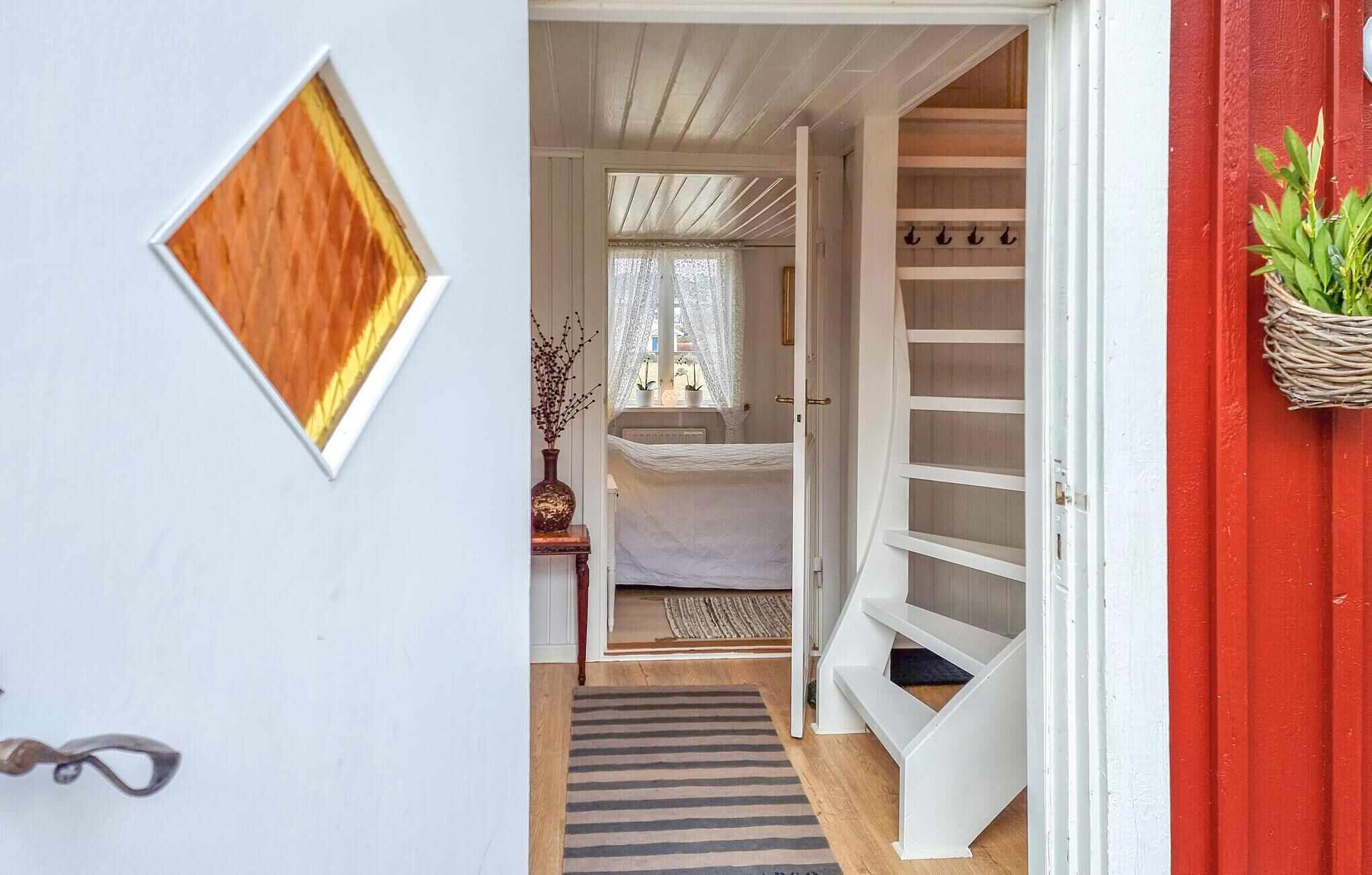 En smal hall leder in till ett sovrum med en säng, ett fönster med spetsgardiner, ett litet bord med en vas och en röd vägg med en växt.