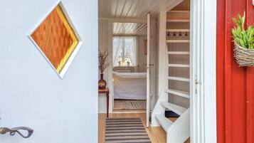 En smal hall leder in till ett sovrum med en säng, ett fönster med spetsgardiner, ett litet bord med en vas och en röd vägg med en växt.