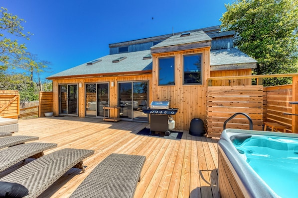 Une terrasse en bois avec des chaises longues, un jacuzzi et un espace barbecue.