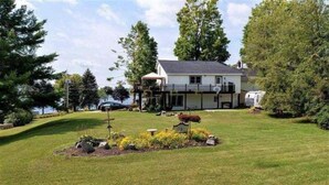 A house with a porch, a garden with plants and a bench, and a boat docked near the water. - Lakeside Retreat with Gorgeous Views Year Round (Schroon Lake)