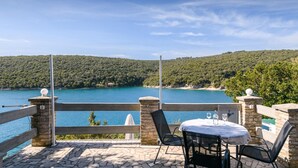 Une terrasse avec un sol en pierre, une table et des chaises, et une vue sur une mer turquoise et des collines verdoyantes.