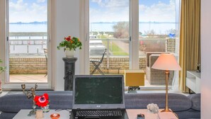 Ett rum med en bärbar dator på ett bord, en vas med blommor, en lampa och utsikt över en vattenyta genom stora fönster.