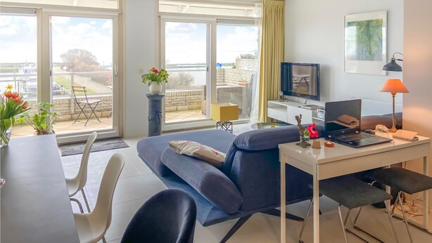 Ett modernt vardagsrum med en soffa, ett matbord och stolar. Det finns en tv, en bärbar dator och en lampa på ett sidobord. Rummet har stora fönster med utsikt över omgivningen.
