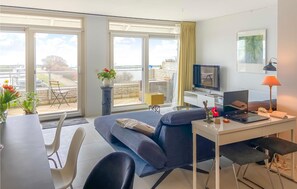 Ein modernes Wohnzimmer mit einer Couch, einem Esstisch und Stühlen. Ein Fernseher, ein Laptop und eine Lampe stehen auf einem Beistelltisch. Große Fenster bieten einen Blick nach draußen.