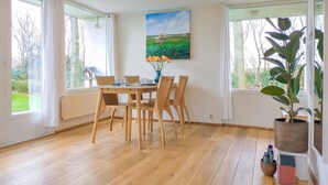 A dining area with a wooden table and chairs, a painting of a landscape, and large windows with curtains.