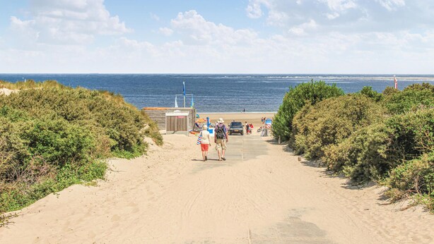 A sandy beach path leading to the ocean with a small building and a flagpole.