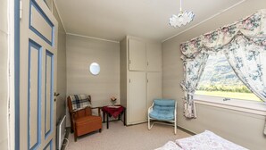 Ein Schlafzimmer mit einem Bett, einem Sessel, einem Kleiderschrank, einem kleinen Tisch mit einer Vase und einem Fenster mit Vorhängen.