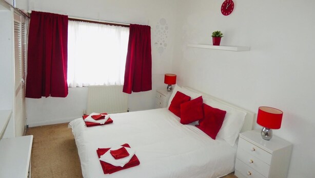 A bedroom with a white bed, red curtains, a red clock, and a red lamp on the nightstand. - Clean & Cosy 3 Bed House with Free Pvt Parking (Croydon)