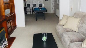A living room with a black coffee table, a beige sofa, a wooden bookshelf, a dartboard, and a ceiling fan. - Clean & Cosy 3 Bed House with Free Pvt Parking (Croydon)