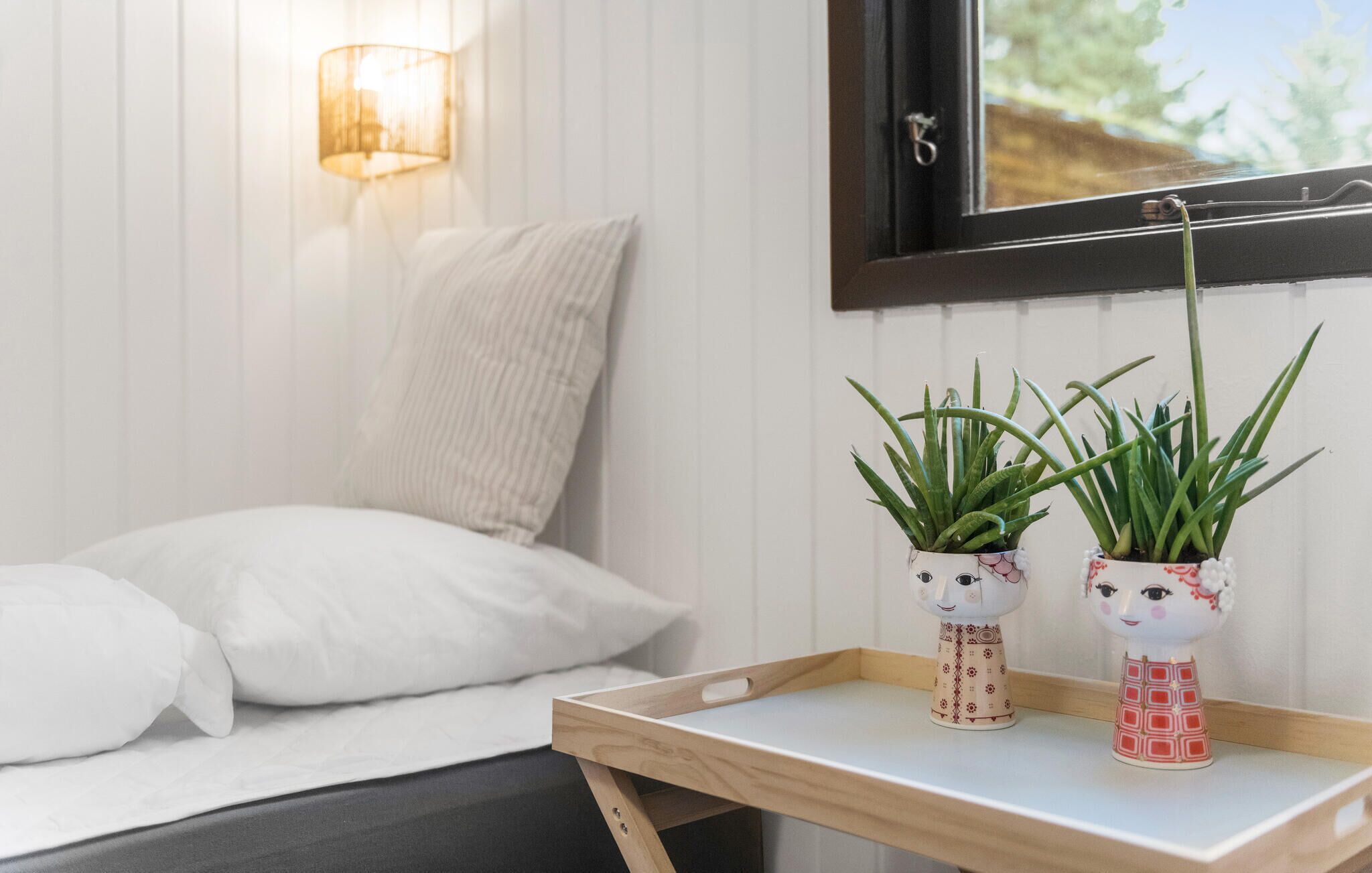 A bed with white bedding, a wooden bedside table with two potted plants, a wall-mounted lamp, and a window with a view of greenery.