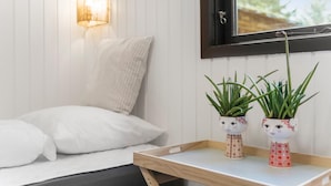 Ein Bett mit weißer Bettwäsche, ein Nachttisch aus Holz mit zwei Blumentöpfen, eine Wandlampe und ein Fenster mit Blick auf Grün.
