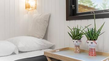A bed with white bedding, a wooden bedside table with two potted plants, a wall-mounted lamp, and a window with a view of greenery.