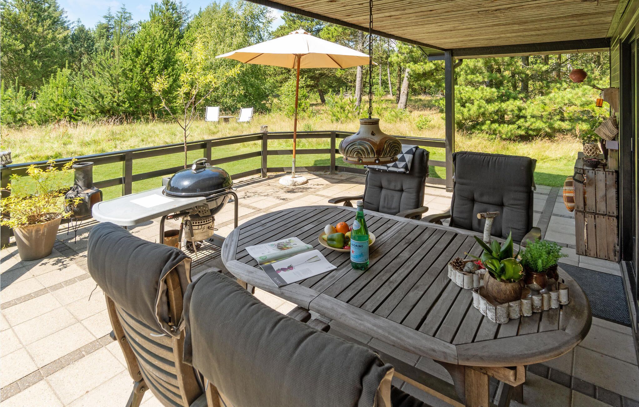 Eine Terrasse im Freien mit Holztisch, Stühlen, einem Grill und einer Pergola mit Sonnenschirm.