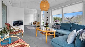A modern living room with a blue sofa, wicker chairs, a wooden coffee table, and a large window with a view of trees. - Stunning home in Ebeltoft with kitchen (Ebeltoft)