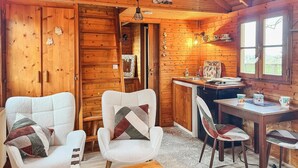A cozy cabin interior with wooden walls, a dining area with a table and chairs, and a kitchen visible through an open door.
