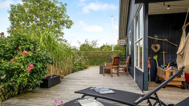 A wooden deck with lounge chairs and a table, surrounded by lush greenery and a building with large windows. - Lovely home in Juelsminde with kitchen (Juelsminde)