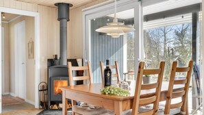 A cozy room with a wood-burning stove, a dining table with chairs, and a large window offering a view of trees. - Lovely home in Juelsminde with kitchen (Juelsminde)