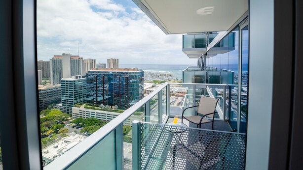 A balcony with a chair and a glass railing overlooking a cityscape with modern buildings and a clear sky.